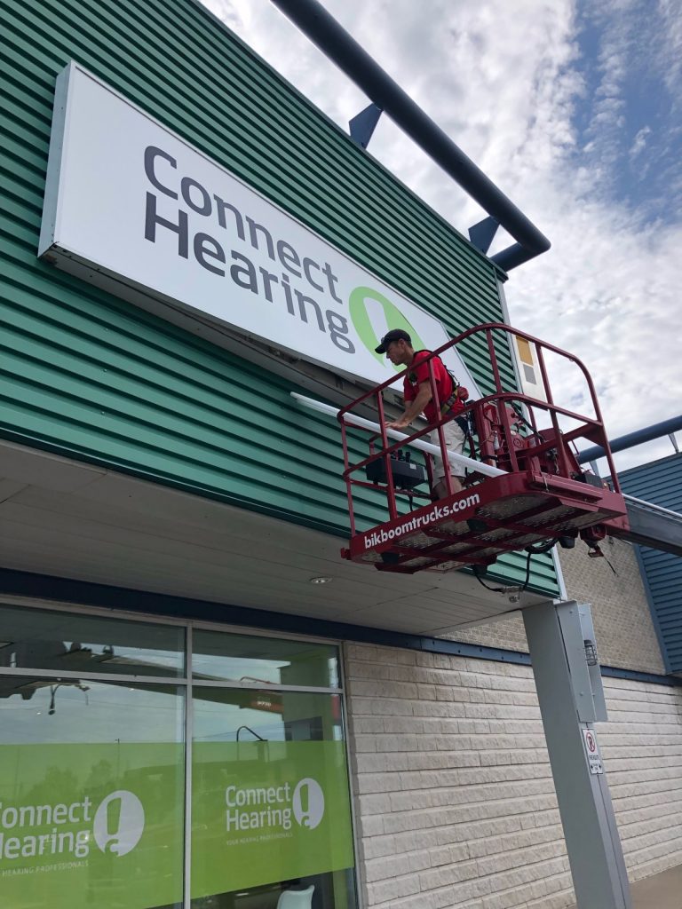 YESCO bucket ladder servicing green and white Connect Hearing sign that is attached to the side of the store