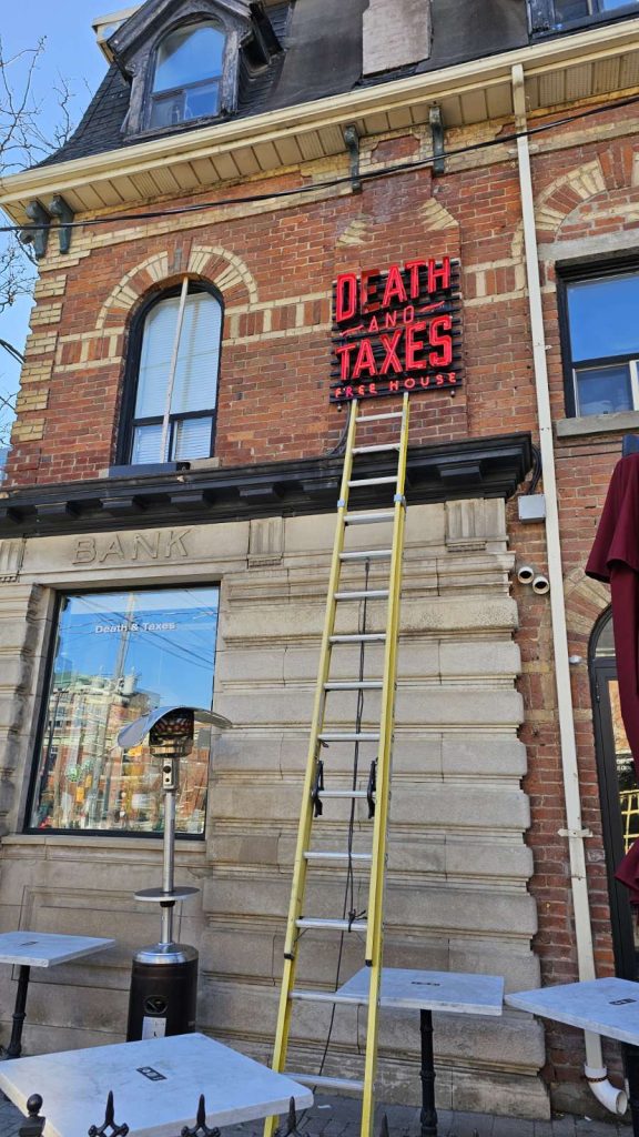 a yellow ladder leaning against an old brick building with a sign saying Death and Taxes.