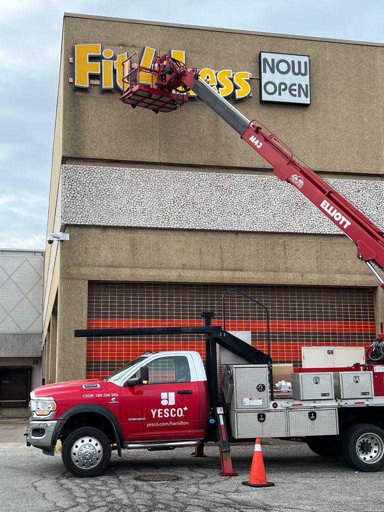 Yesco truck with bucket ladder at the top of a building servicing the Fit $ Less sign