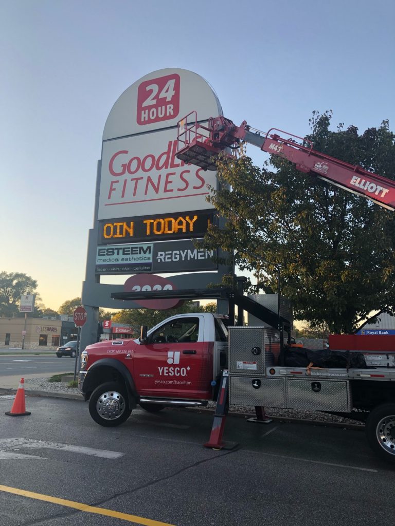 Yesco truck with bucket ladder servicing the red and white lettered GoodLife Fitness sign