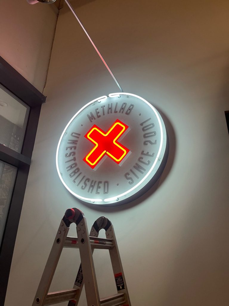 a neon sign with a red neon cross in the centre and the words methlab unestablished since 2001