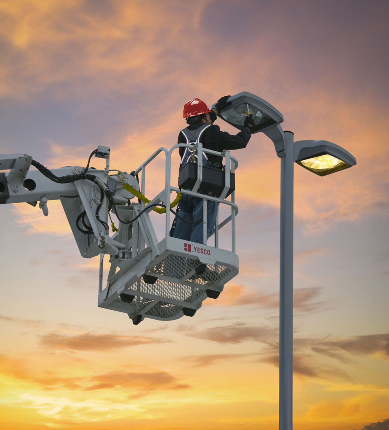 A worker wearing a red helmet and safety harness stands on a lift, repairing a streetlight at sunset.