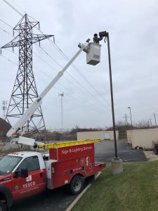 a photo of a Yesco truck and employee in a bucket ladder fixing a pole light in a parking lot