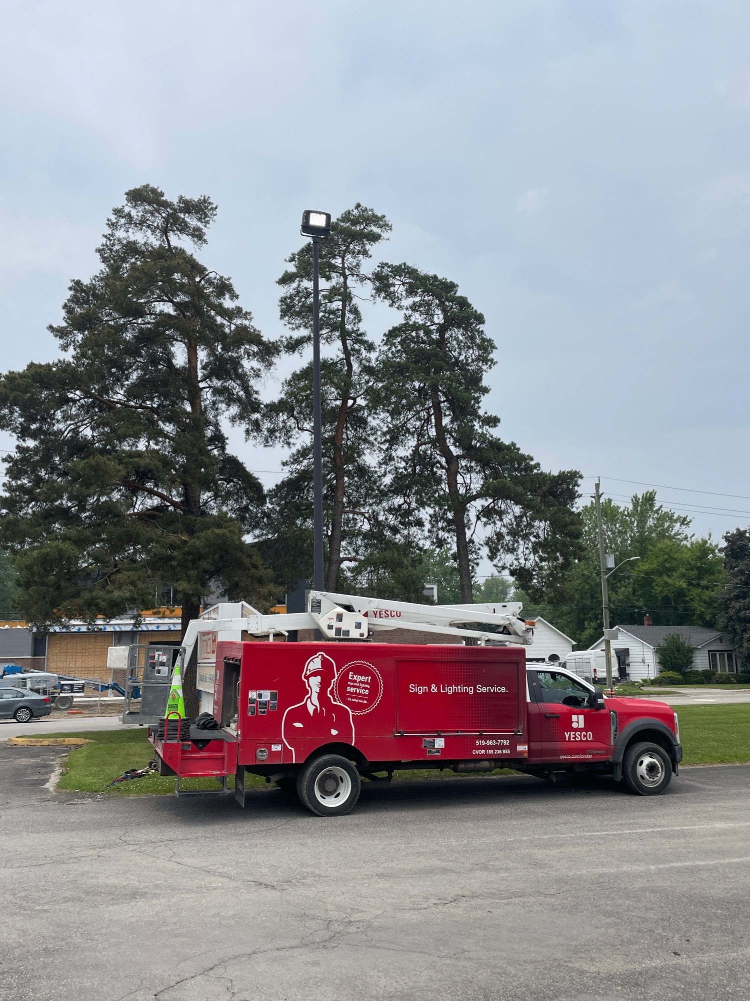 a red Yesco service truck parked in a parking lot with a tall poll light behind it and tall trees in the background
