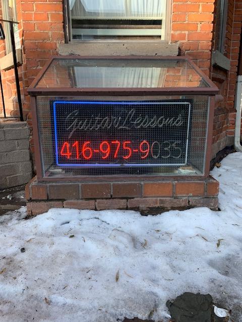 red and blue neon sign repair on small sign at front of building that says Guitar Lessons 416-975-9035
